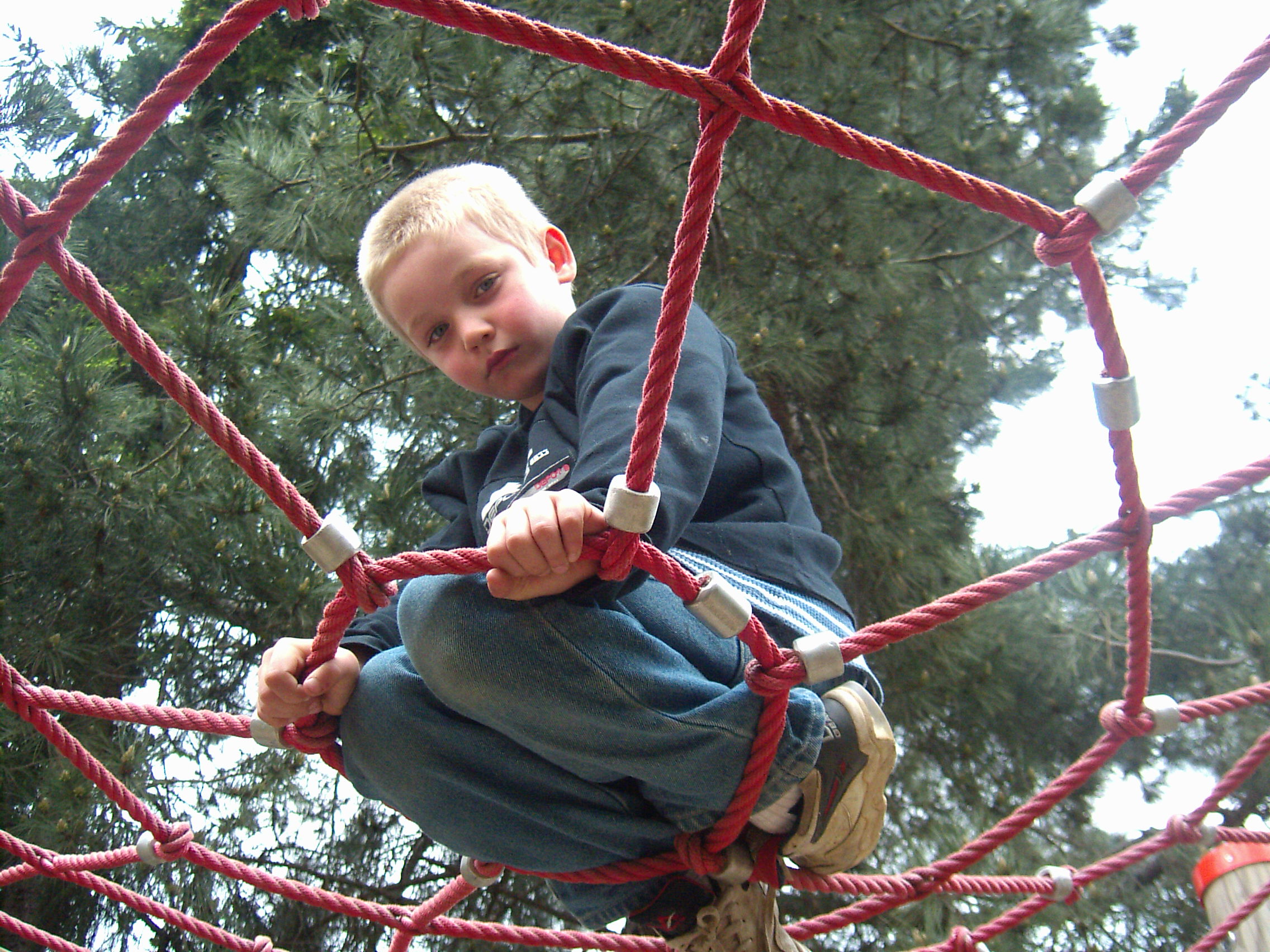 zu sehen ist ein von unten fotografierter kleiner Junge, der knieend von einem Spinnennetz eines Kinderspielplatzes gehalten wird, und in die Kamera schaut